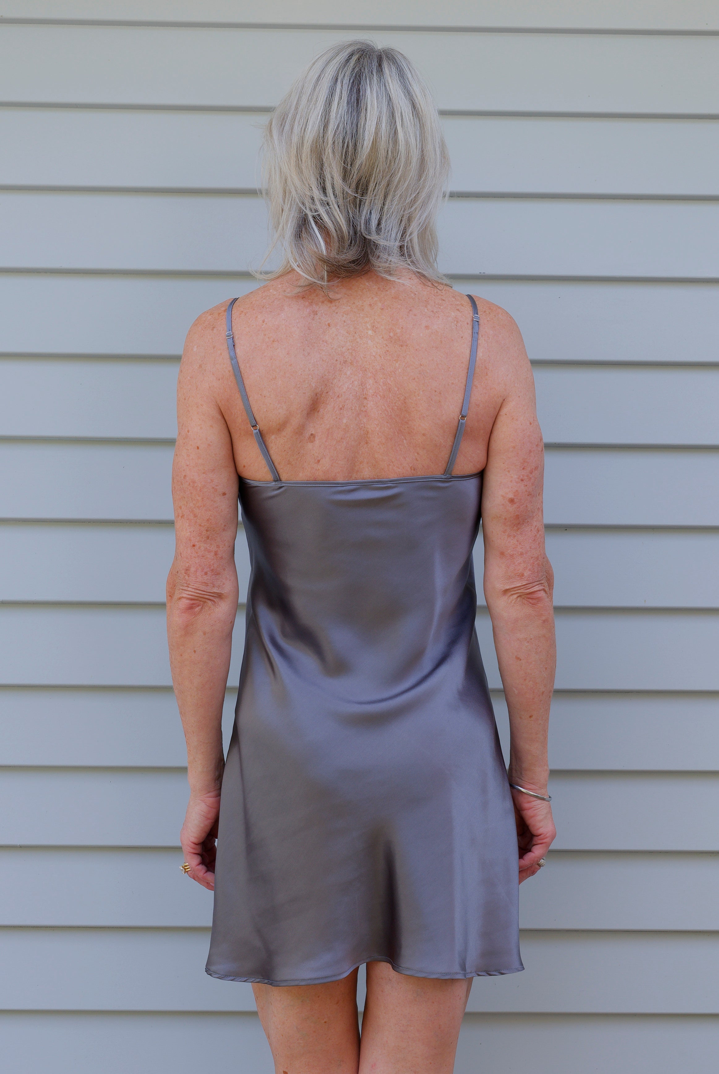 Woman wearing a grey silk slip dress standing against a grey wall.
