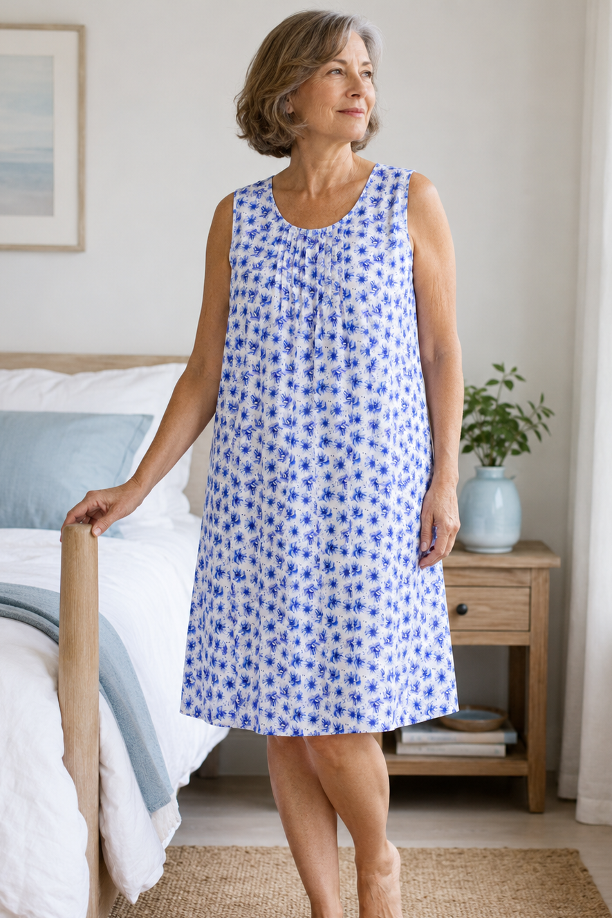 An older woman stands barefoot in a softly lit bedroom wearing a sleeveless knee-length nightgown with a blue floral print on a white background. The room includes a bed, wooden nightstand, plant, and window with white curtains.