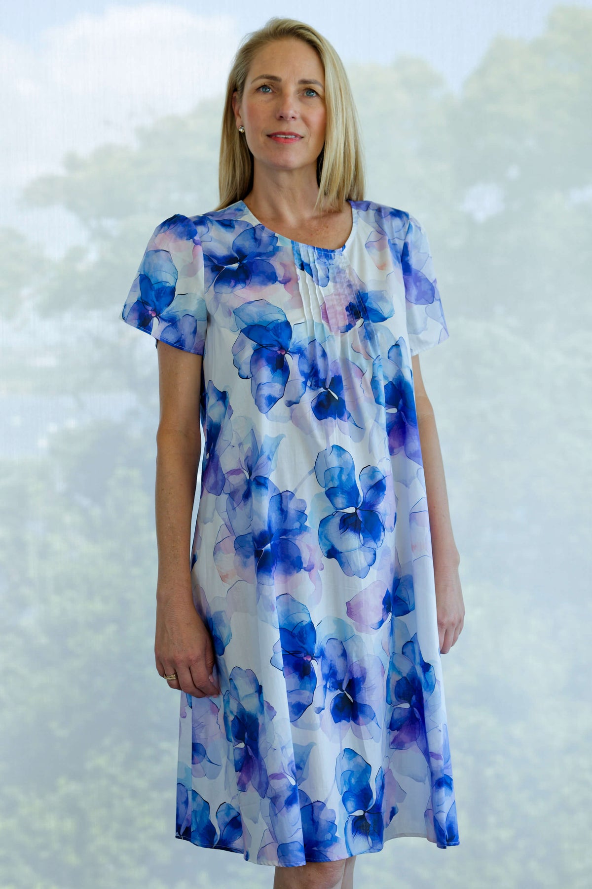 A woman modeling a short-sleeve midi cotton nightie with a blue and purple watercolor floral print on a white background, set against a blurred green forest backdrop.