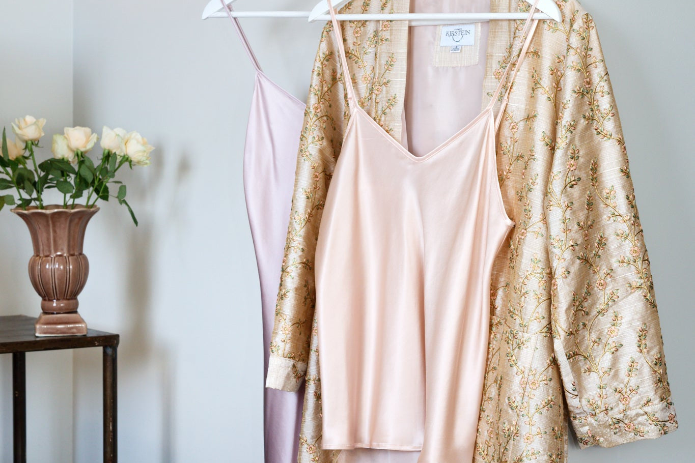 silk sleepwear items on hangers against a wall with a vase of flowers on a table.