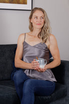 A woman sitting on a couch wearing a gray silk camisole top paired with blue jeans, holding a cup.