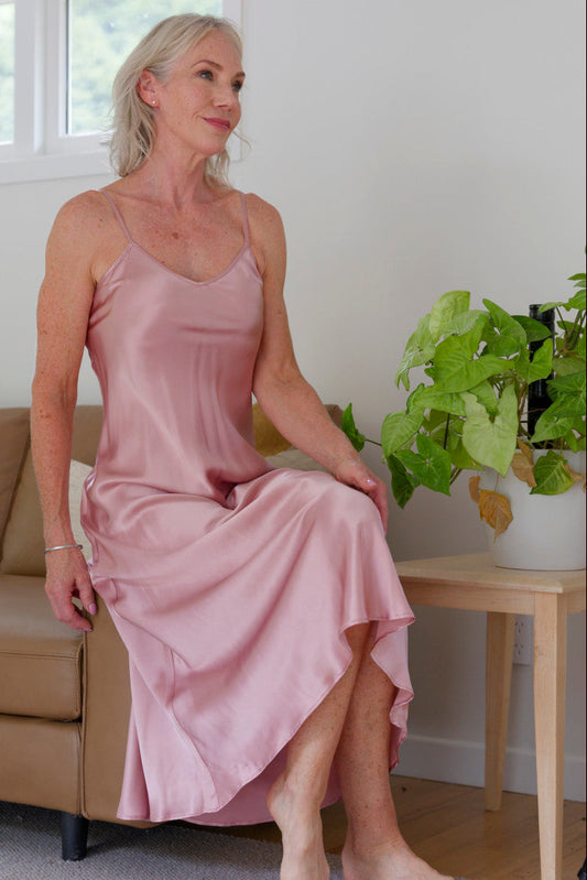 Woman in a pink silk satin night dress sitting on a couch in a room with a plant.