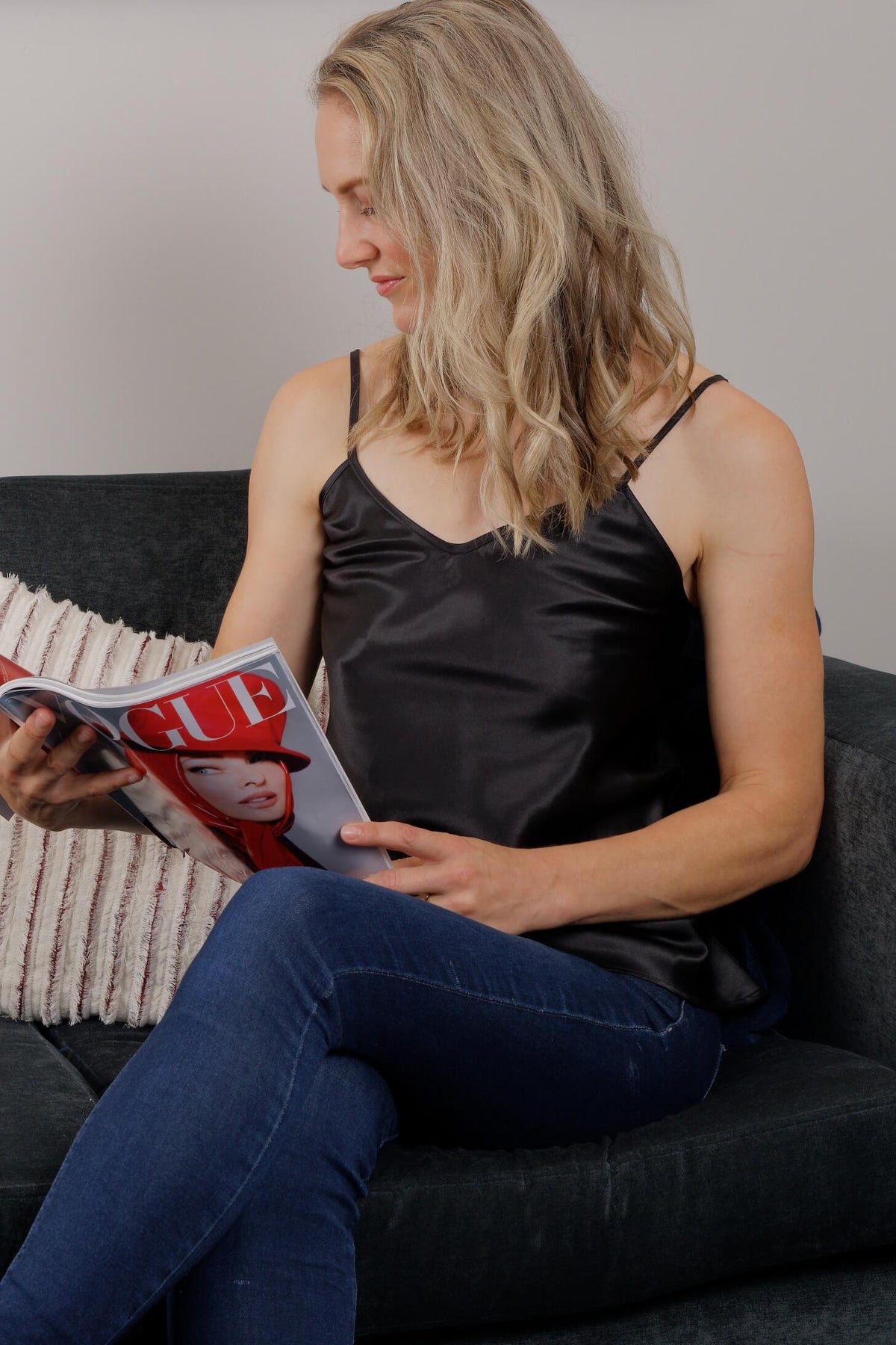 A woman with blonde hair sits on a dark gray couch, wearing a black satin camisole top and blue jeans while holding a magazine.