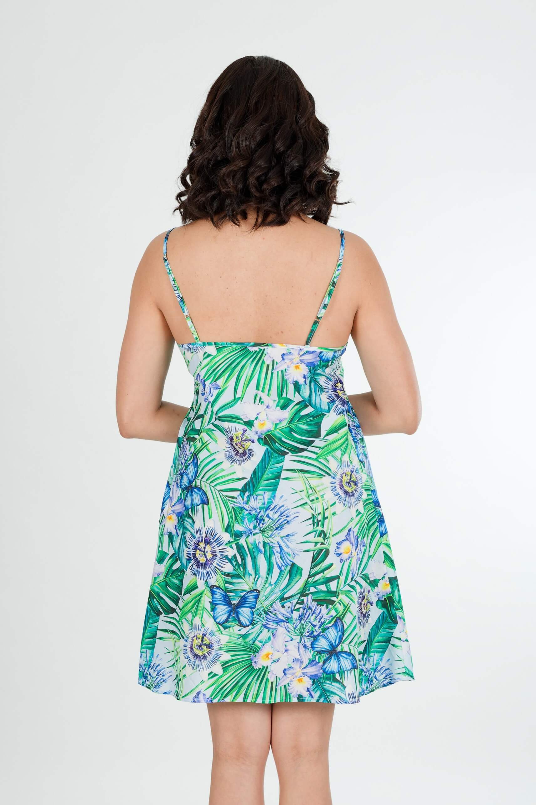 A woman viewed from the back wearing a sleeveless tropical print mini dress with thin straps and a flared skirt, set against a plain light gray studio background.
