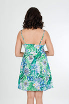 A woman viewed from the back wearing a sleeveless tropical print mini dress with thin straps and a flared skirt, set against a plain light gray studio background.