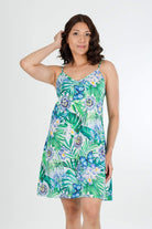 A woman modeling a sleeveless slip-style dress with a tropical floral and leaf print in shades of green, blue, and white, set against a plain light gray studio background.