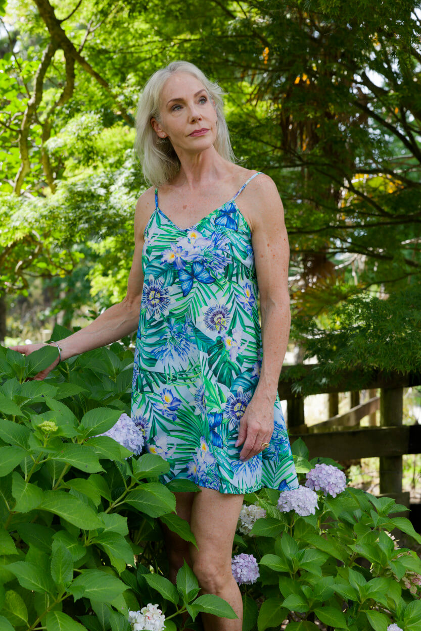 A woman wearing a green and blue tropical floral print slip dress stands outdoors in a garden with green foliage and purple flowers.