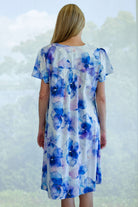 A woman viewed from behind wearing a short-sleeve cotton night dress with a blue and purple watercolor floral print, set against a soft-focus outdoor background.