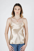 A woman modeling a shiny champagne-gold satin camisole top with blue jeans, standing against a light gray studio background.