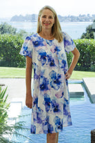 A woman wearing a cotton short-sleeve white nightdress with a blue and purple watercolor floral print, standing in front of a swimming pool and green garden background.